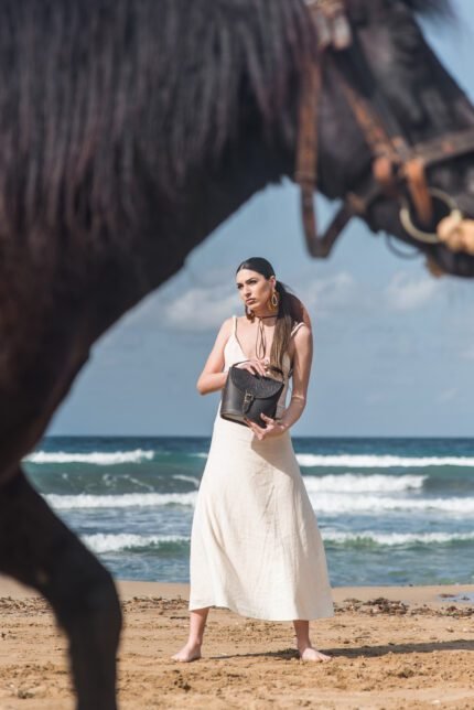Woman standing barefoot on the beach in a white dress holding a Jirakis Handcrafted Leather Embossed Shoulder Bag for women, with a horse in the foreground.