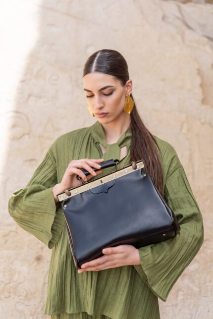 Model showcasing a glossy black leather doctor bag with metal hardware, wearing a green dress outdoors