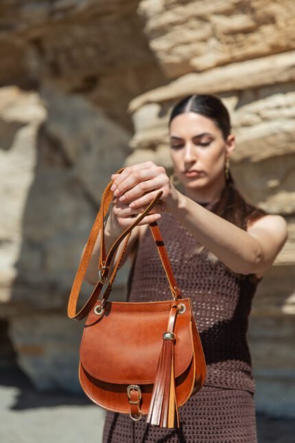 Woman holding a brown leather crossbody saddle bag with buckle and tassel detail