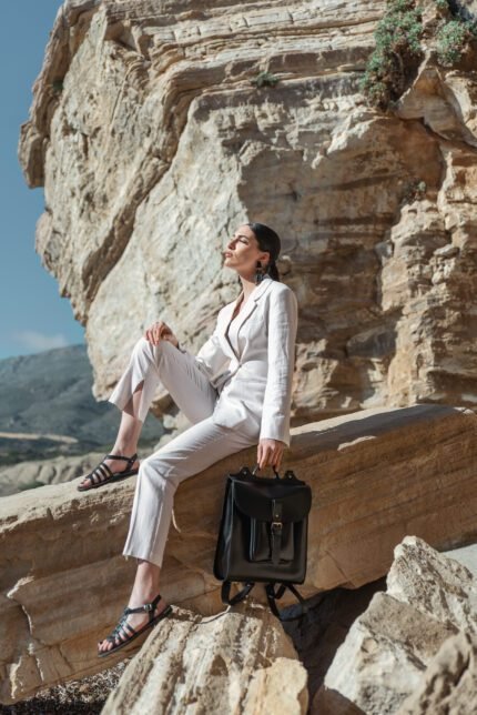 Woman in white suit sitting on rock with black leather flap backpack by her side, handcrafted by Jirakis Handcrafted Leather in Crete.