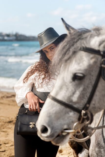 Woman with black leather lock satchel bag standing on the beach beside a white horse – handcrafted Greek leather bag