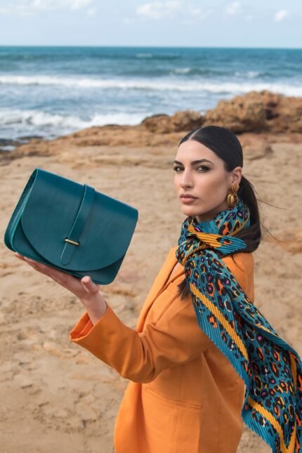Model holding a teal leather everyday bag with flap closure, styled with a scarf and orange coat, Jirakis Handcrafted Leather.