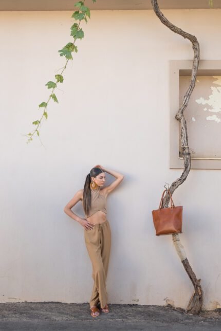 Woman in beige outfit posing next to a brown leather tote bag hanging on a vine against a white wall