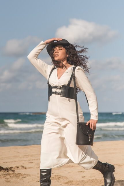 Woman walking on the beach wearing a white dress, black boots, and carrying a Jirakis Handcrafted Leather Embossed Crossbody Bag for women.
