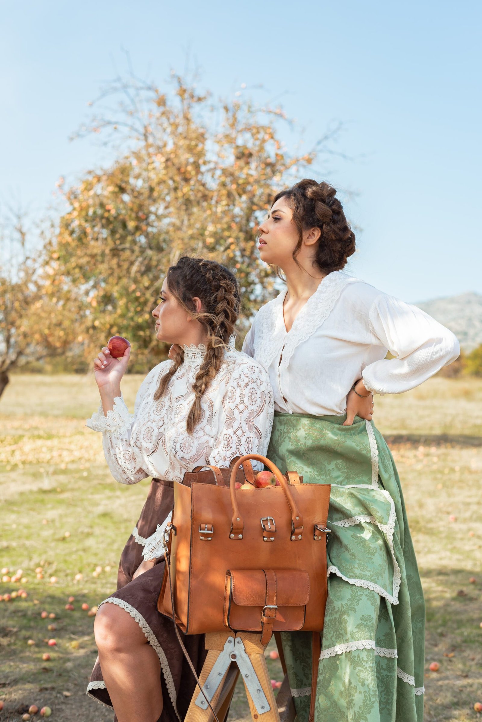 Brown Spartan Nomad Leather Carryall Bag – Jirakis Handcrafted Leather Two women with brown Spartan Nomad leather carryall bag with buckles, sitting in orchard, handcrafted in Crete by Jirakis Handcrafted Leather.