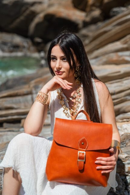 Model wearing an orange leather modern backpack with buckle, handcrafted by Jirakis Handcrafted Leather by the sea.