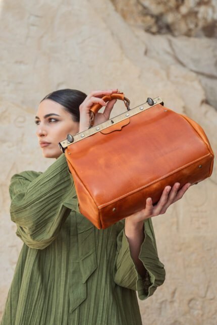 Woman holding a large tan handmade leather travel bag for women in vintage doctor-style design, photographed against a stone wall in Crete.