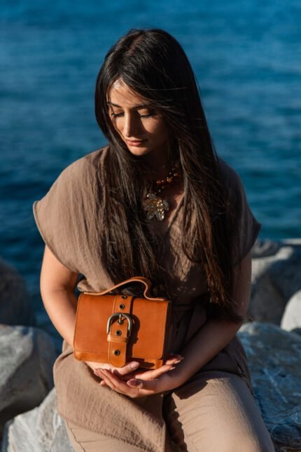Woman holding small structured leather mini satchel with buckle detail, handcrafted by Jirakis Handcrafted Leather in Crete.