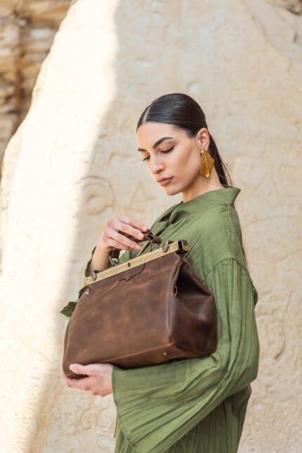 Woman in green dress showcasing a brown leather case with vintage brass hardware against a carved stone background