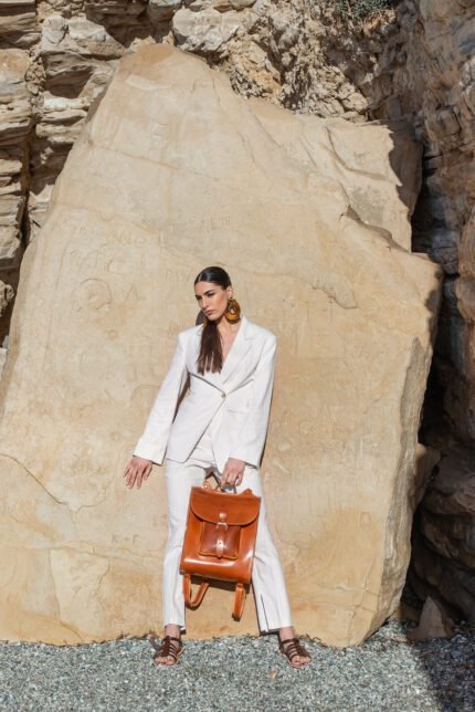 Woman in white suit holding brown leather backpack with buckle front pocket, handcrafted by Jirakis Handcrafted Leather in Crete.